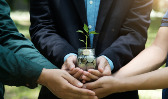 Hands of three people holding a glass jar of money with a plant growing within it