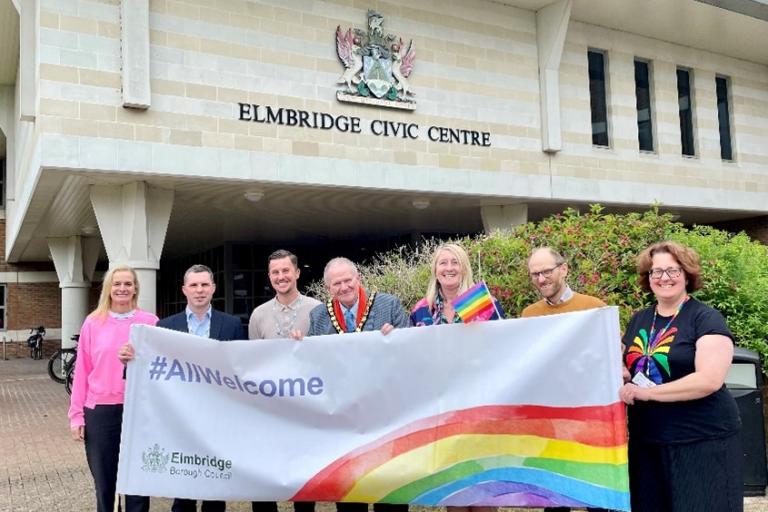 Seven people, including the mayor, holding a banner featuring a rainbow and 'All Welcome' message in front of the Elmbridge Civic Centre