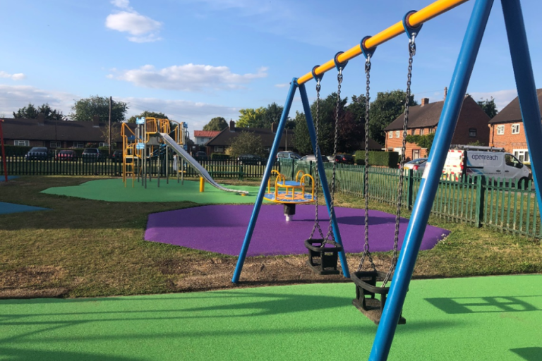 Children's playground featuring a blue and orange swing set in the foreground and slide in the background with bright green and purple flooring.