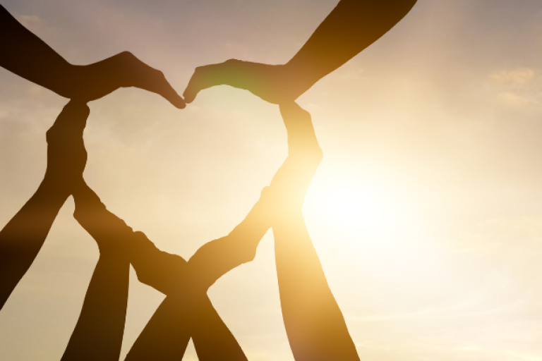 Hands coming together to make a heart shape silhouette in front of the sun.