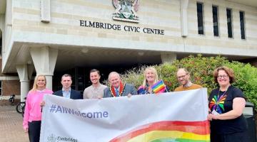 Seven people, including the mayor, holding a banner featuring a rainbow and 'All Welcome' message in front of the Elmbridge Civic Centre