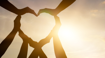 Hands coming together to make a heart shape silhouette in front of the sun.